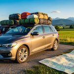 Voiture familiale chargée de bagages sur une aire de départ, avec une carte routière ouverte et un paysage estival de campagne et de montagnes en arrière-plan.