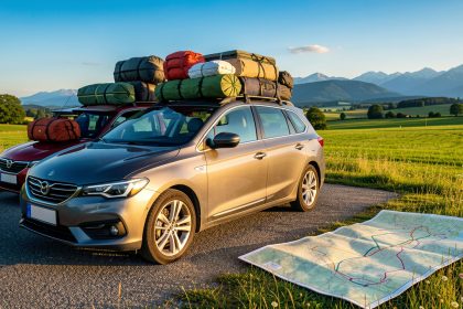 Voiture familiale chargée de bagages sur une aire de départ, avec une carte routière ouverte et un paysage estival de campagne et de montagnes en arrière-plan.