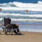 Personne en fauteuil roulant avançant sur un tapis d’accès vers une plage des Antilles.