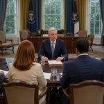 Vue éloignée et réaliste du Bureau ovale avec un président américain assis à une table d’interview face à quatre journalistes vus de dos, dans une lumière sobre.
