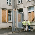 Façade d’un lycée public français après un blocus, avec portail fermé, vitres protégées et deux agents techniques évaluant des dégradations légères.
